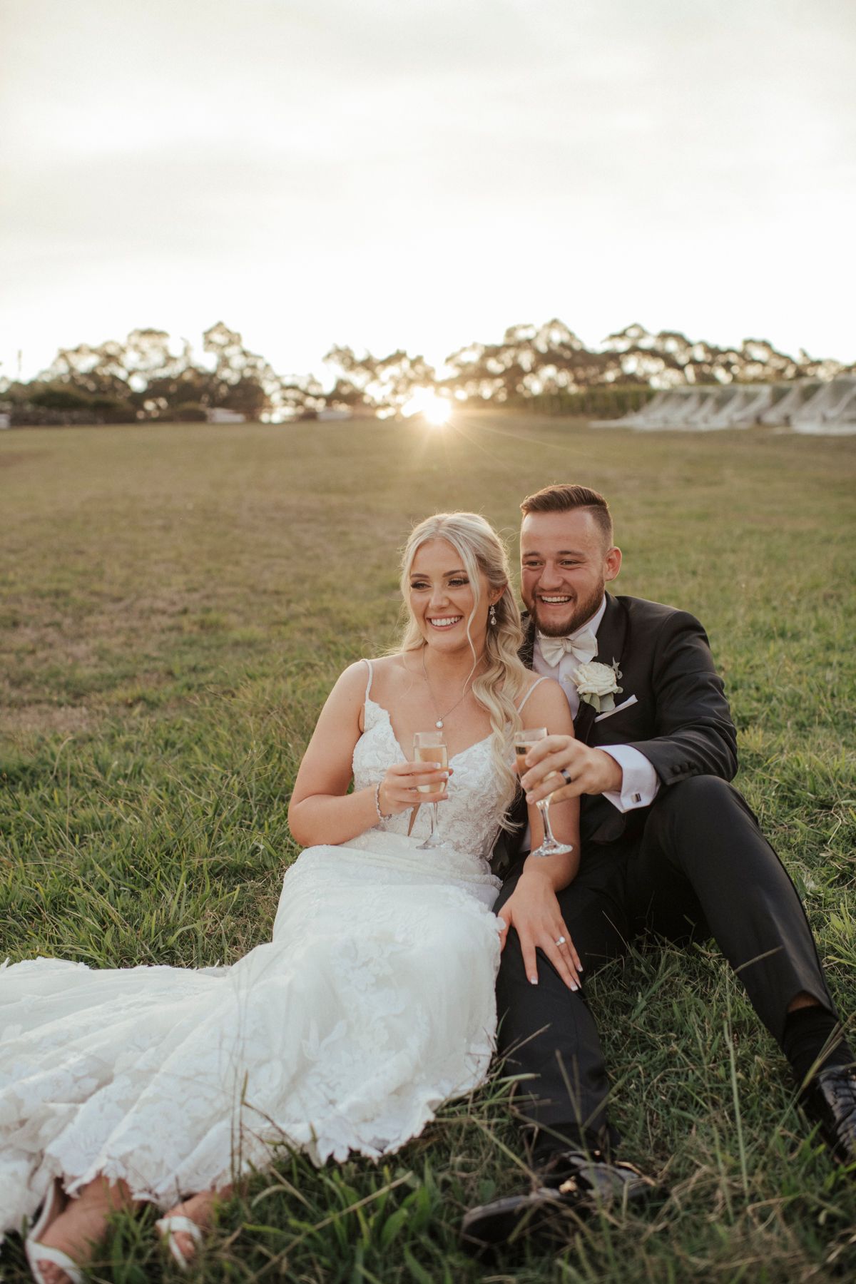 Newlyweds relax in a grassy field at sunset while holding champagne glasses.