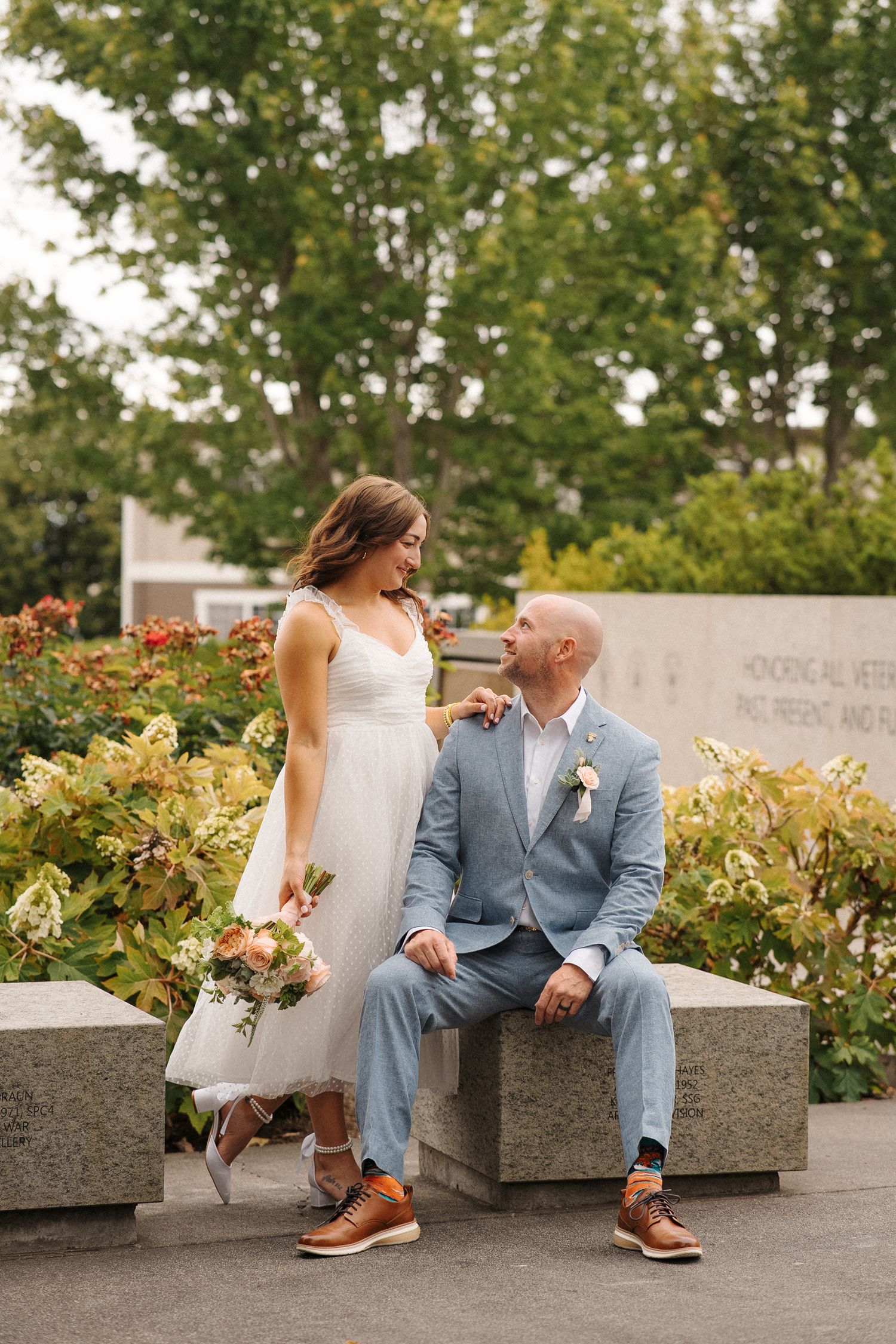 A bride in a white dress and companion in blue suit sitting together on stone steps surrounded by garden flowers.