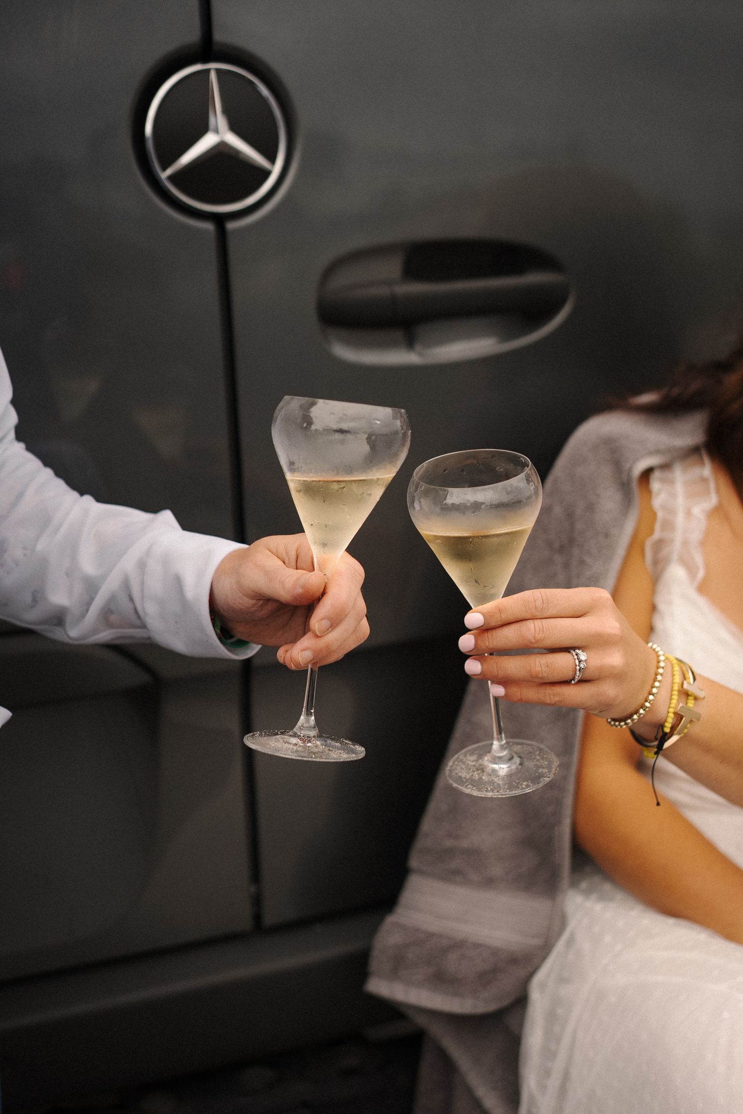 Two hands holding champagne glasses toasting in front of a Mercedes-Benz logo on a dark background.