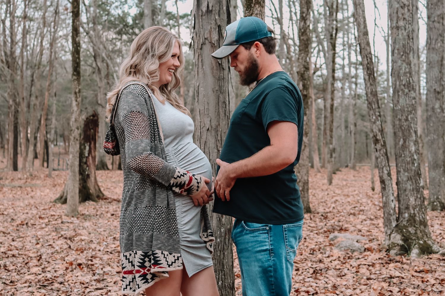 A couple shares an intimate moment in a wooded forest setting surrounded by fallen autumn leaves.