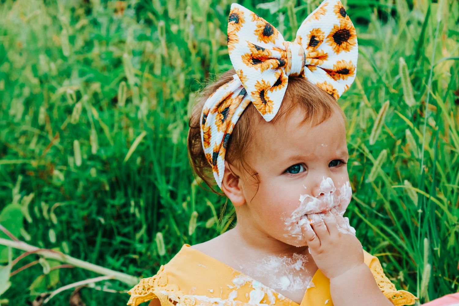 A small child in yellow dress with floral bow headband sits in grass with cake frosting smeared on their face.