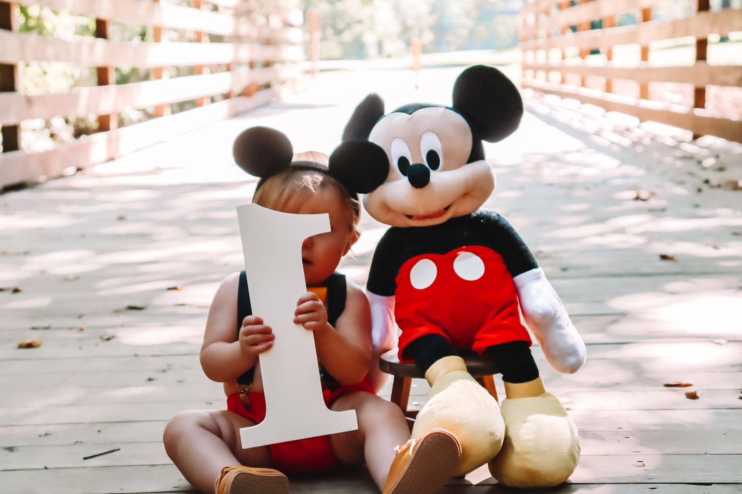 Mickey Mouse plush toy sits next to a wooden number one decoration on wooden bridge.