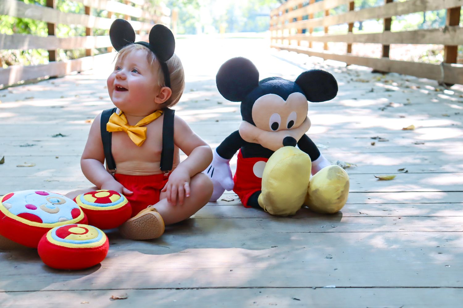 Toddler in Mickey Mouse costume sits on wooden deck next to plush Mickey Mouse toy and plated cookies.
