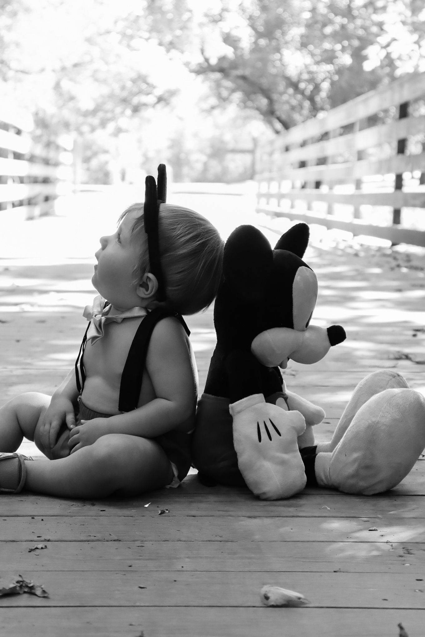 Black and white image of a child sitting back-to-back with a Mickey Mouse plush toy on a wooden deck.