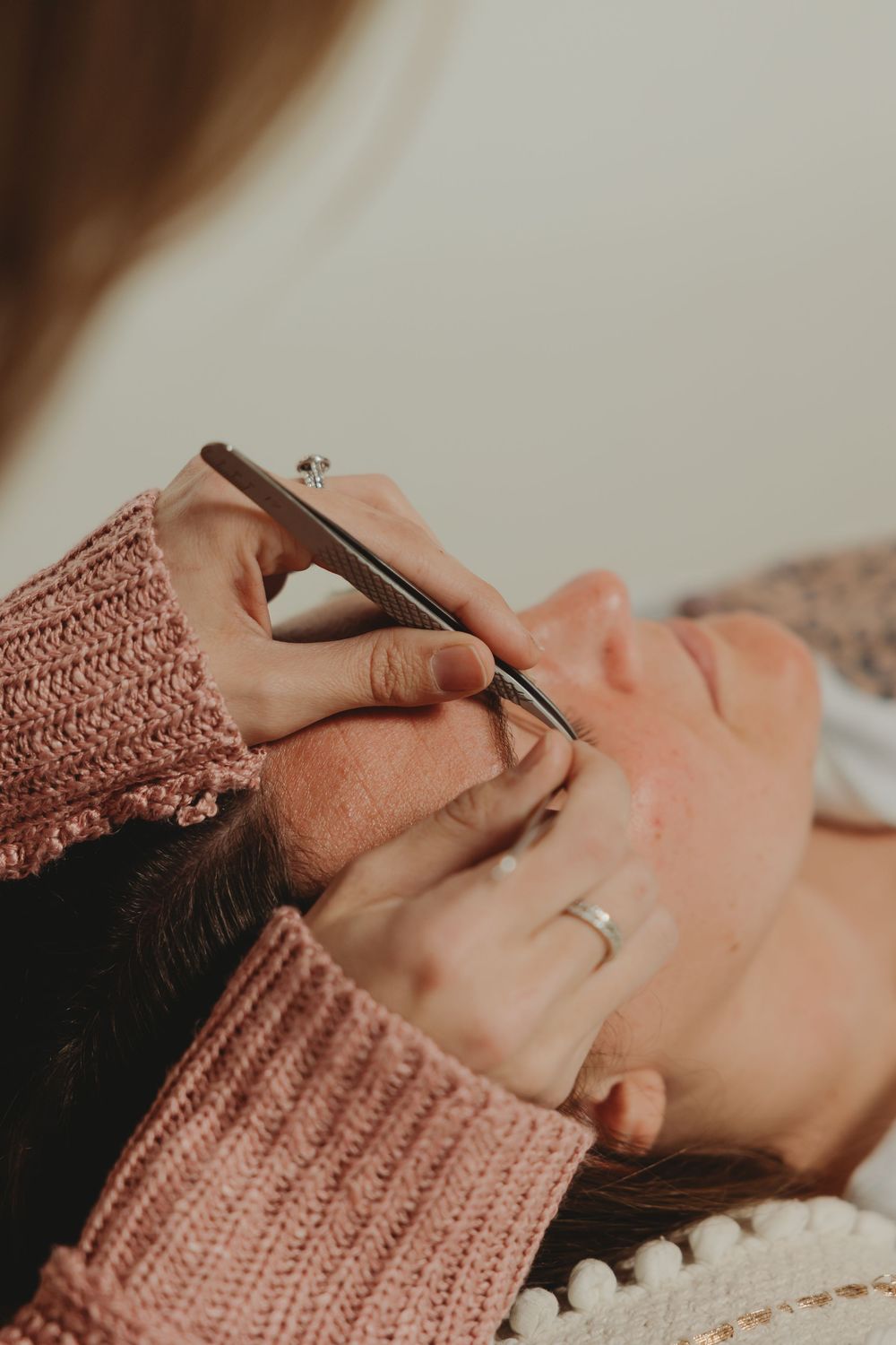 Close-up of hands in pink knit sweater carefully applying individual eyelash extensions during beauty treatment.