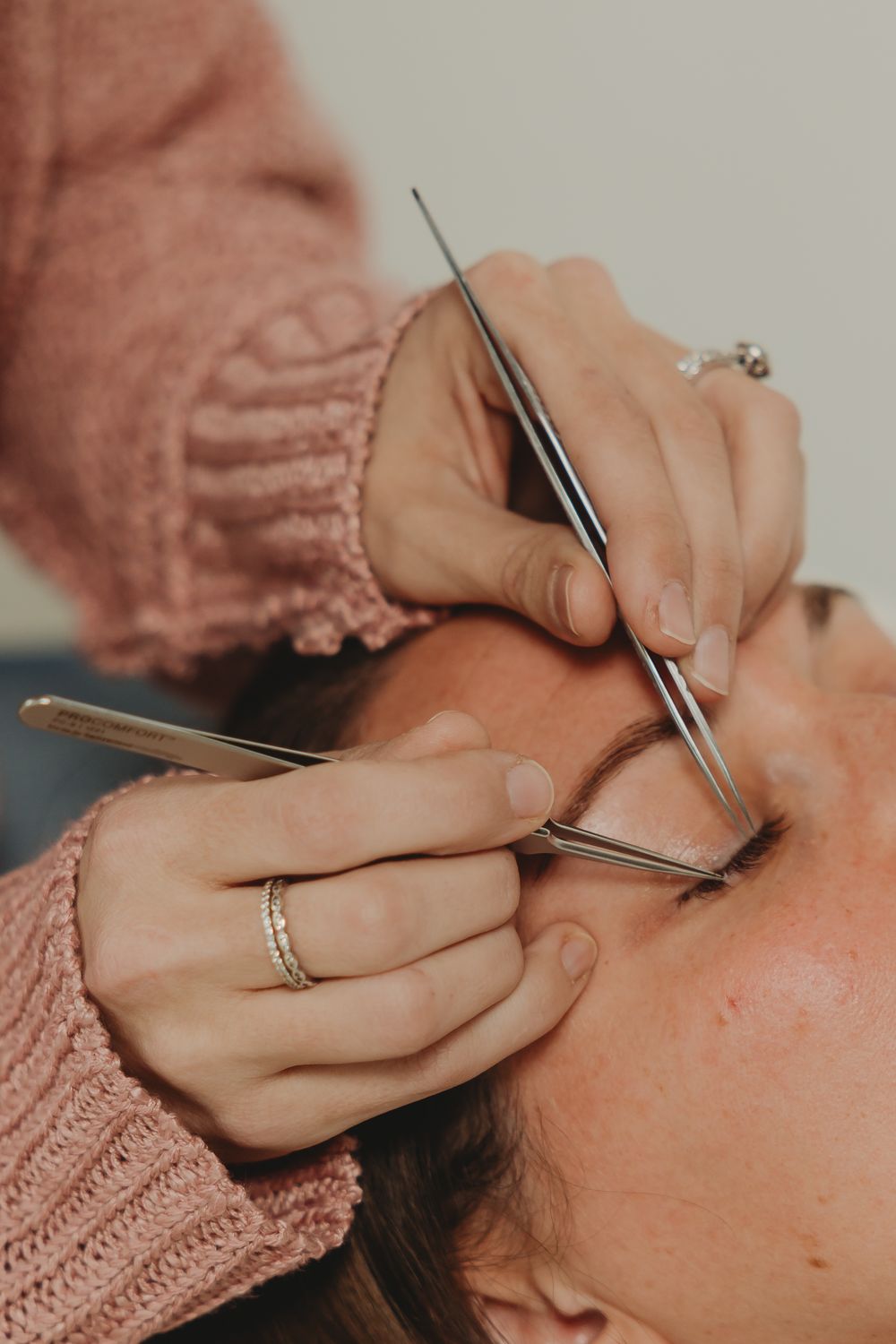 Hands wearing pink sweater work on eyelash extensions with professional tweezers while wearing delicate ring jewelry.
