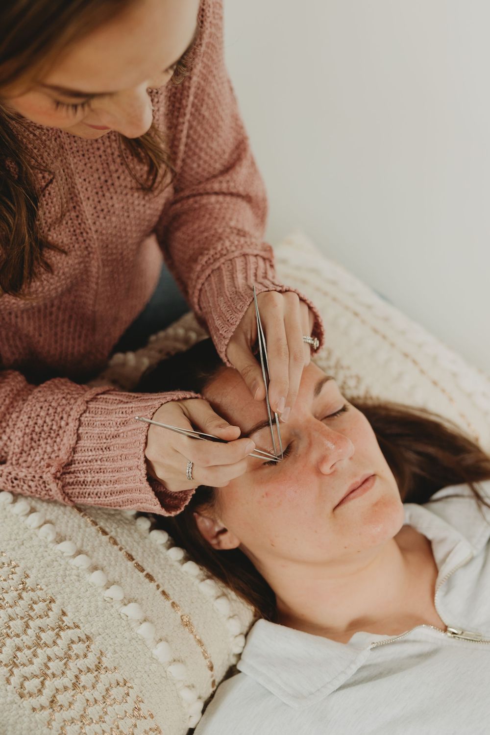 Person in a pink knit sweater performing facial acupuncture treatment on someone laying on a textured blanket.