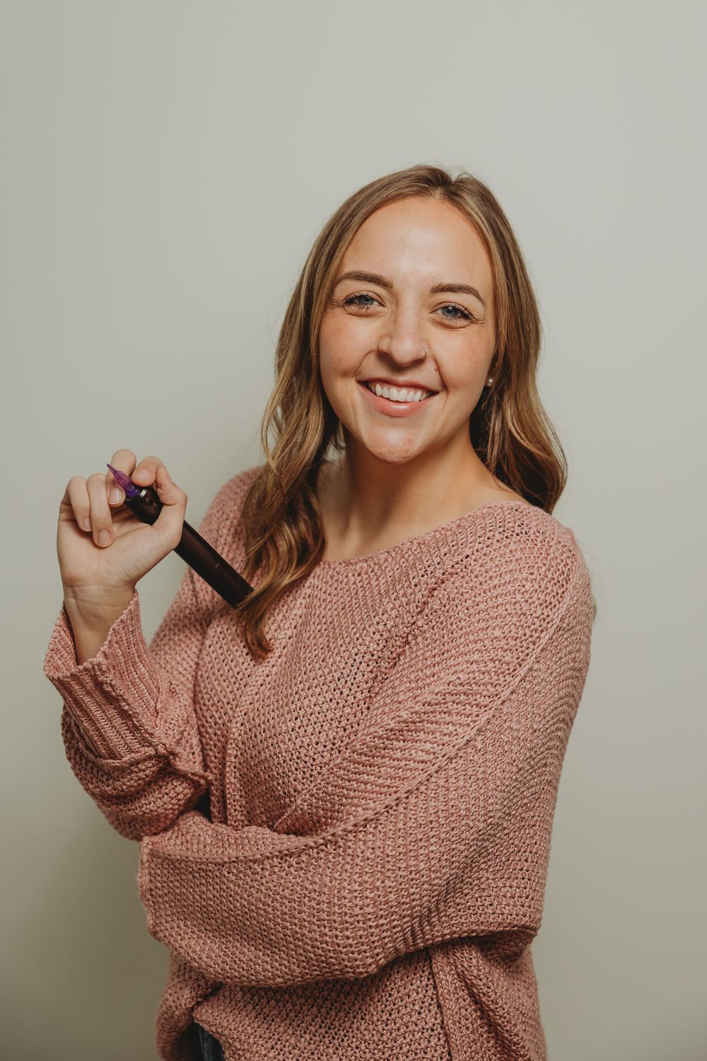 A person in a cozy pink knit sweater smiles warmly while playfully touching their long wavy hair against a neutral backdrop.