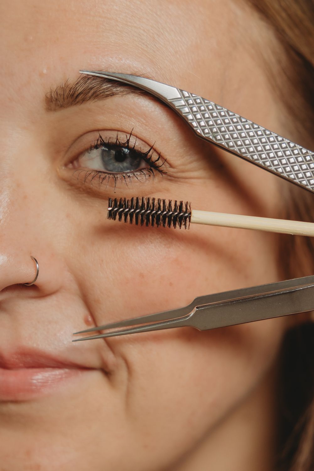 Close-up beauty shot showing eyebrow grooming tools including tweezers, spoolie brush and textured metal shaping tool.