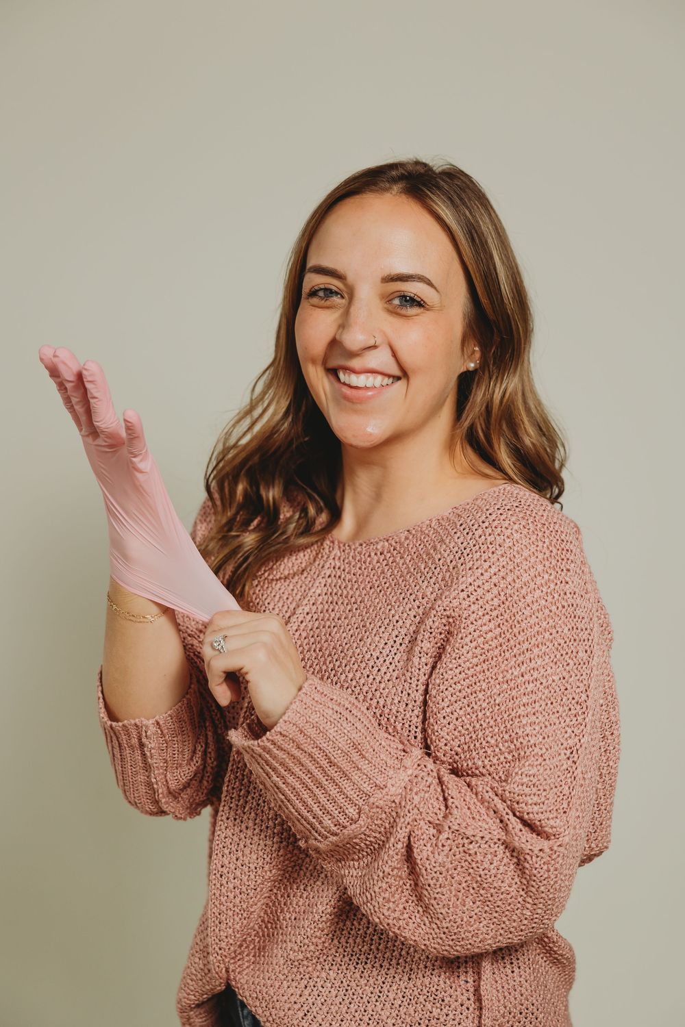 Someone wearing a pink knit sweater smiles while putting on a pink disposable glove against a neutral background.