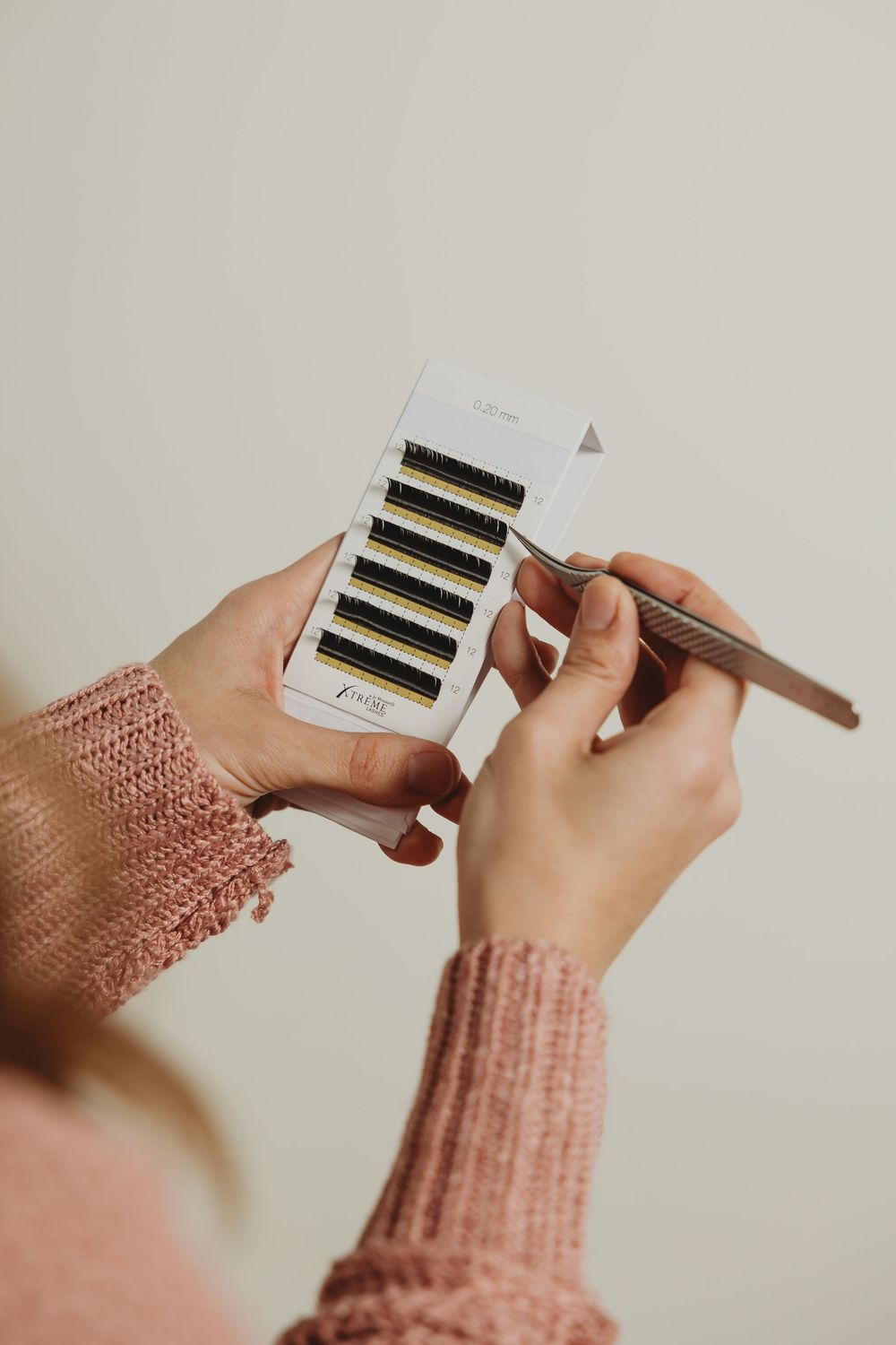 Hands wearing a pink knit sweater hold and open a small white box against a neutral background.