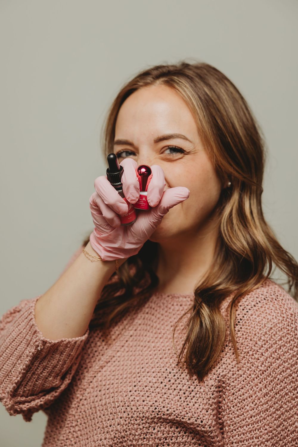 Someone wearing a pink knit sweater smiles while holding a lipstick tube near their face.