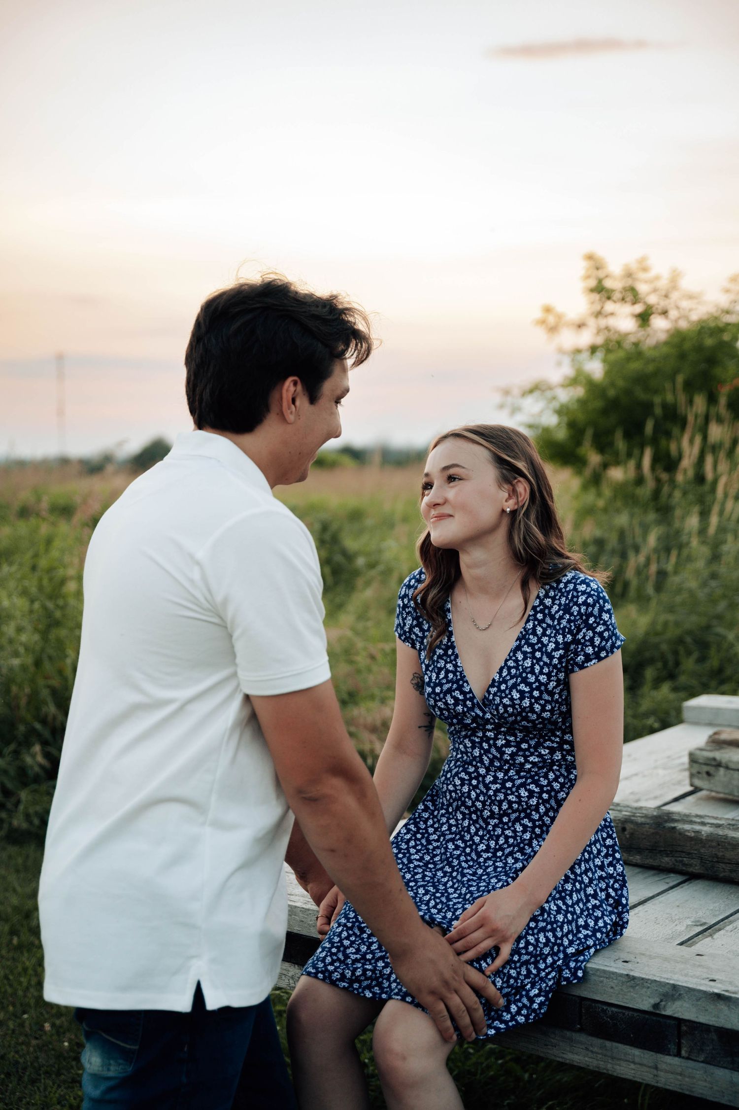 A couple sits together on a rustic bench during sunset, she wearing a blue polka dot dress and he in a white shirt.