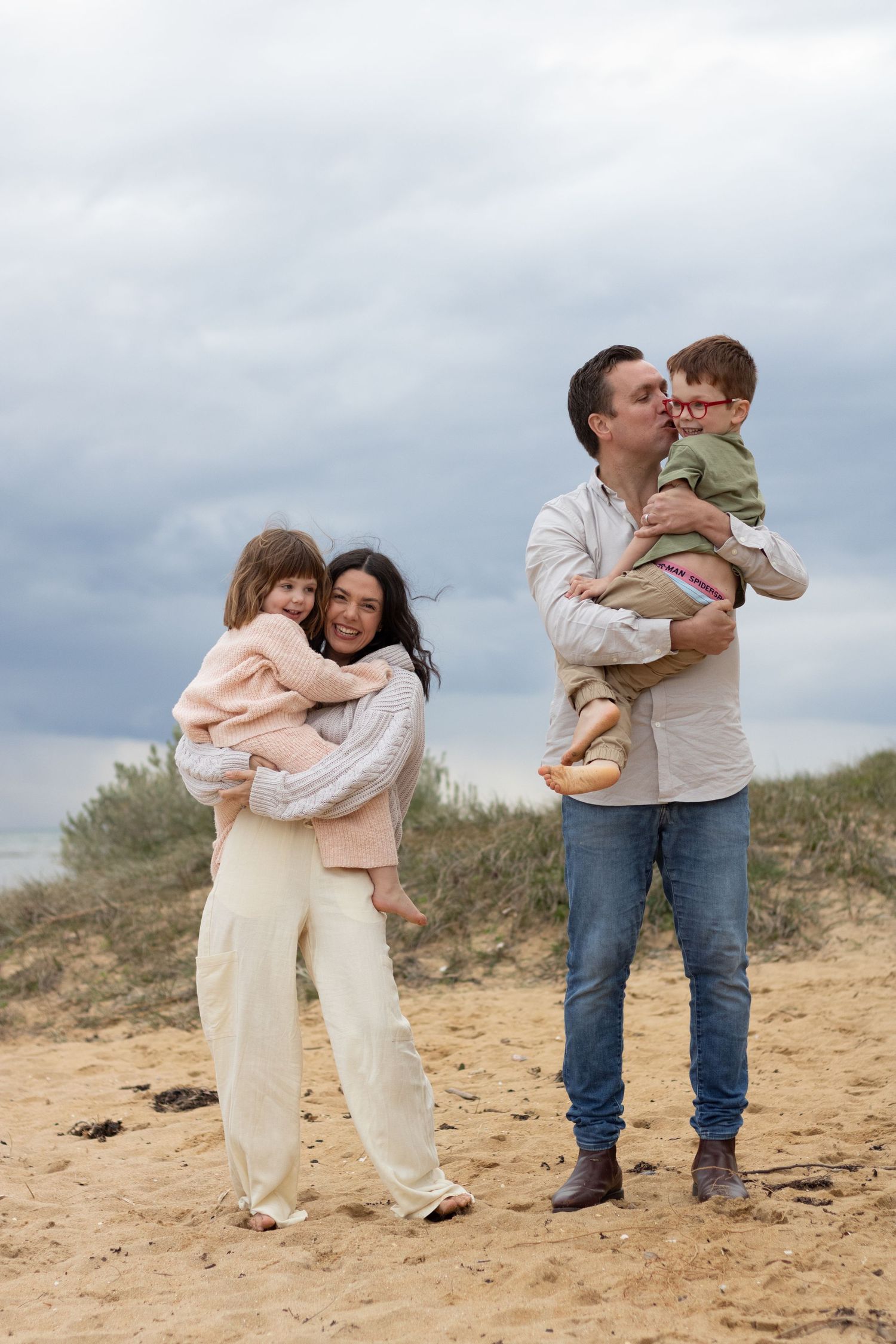 A loving family embraces while standing on a sandy beach against a moody sky backdrop.