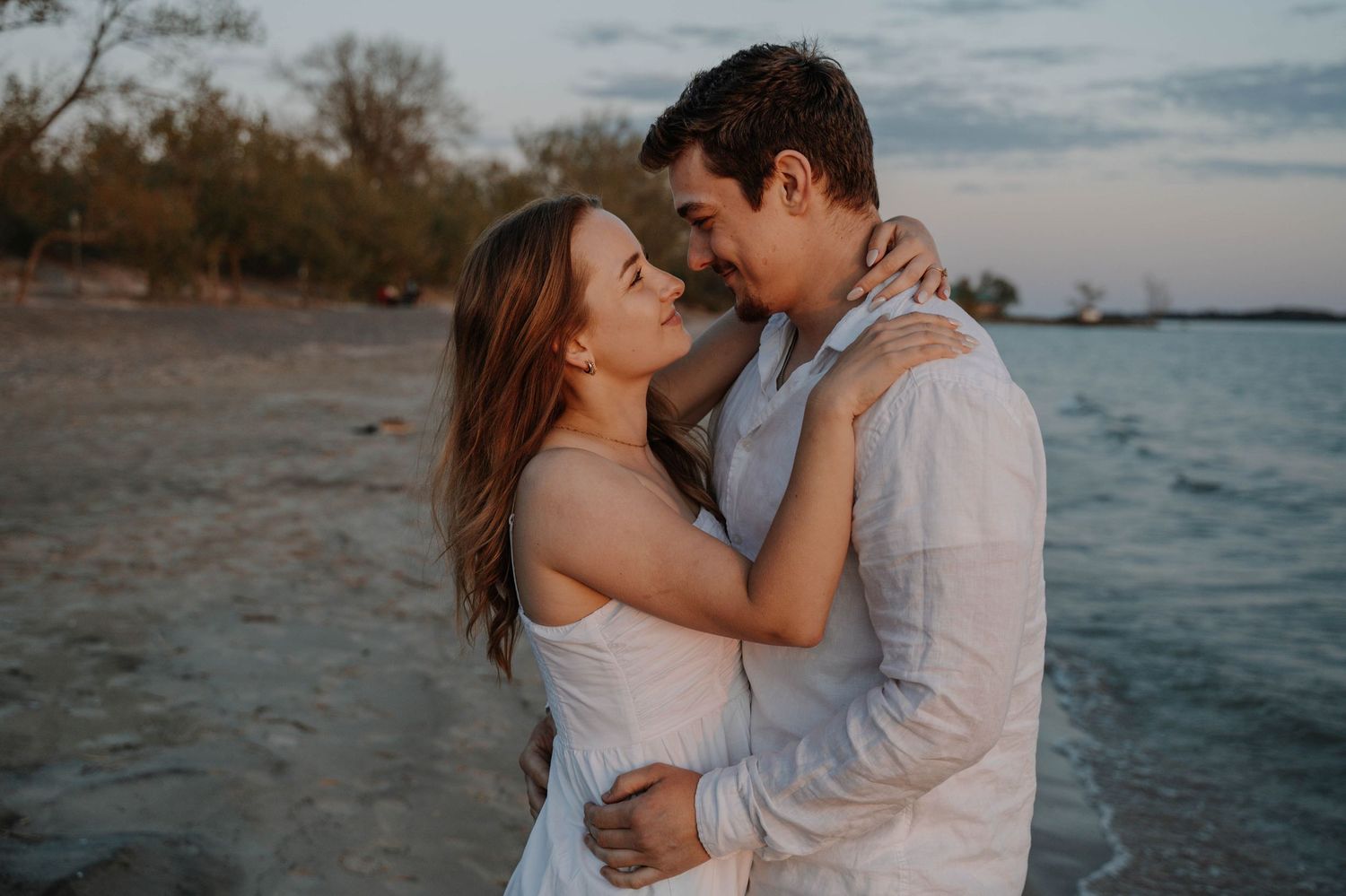 A romantic couple embraces at sunset by a lake shore with trees in the background.