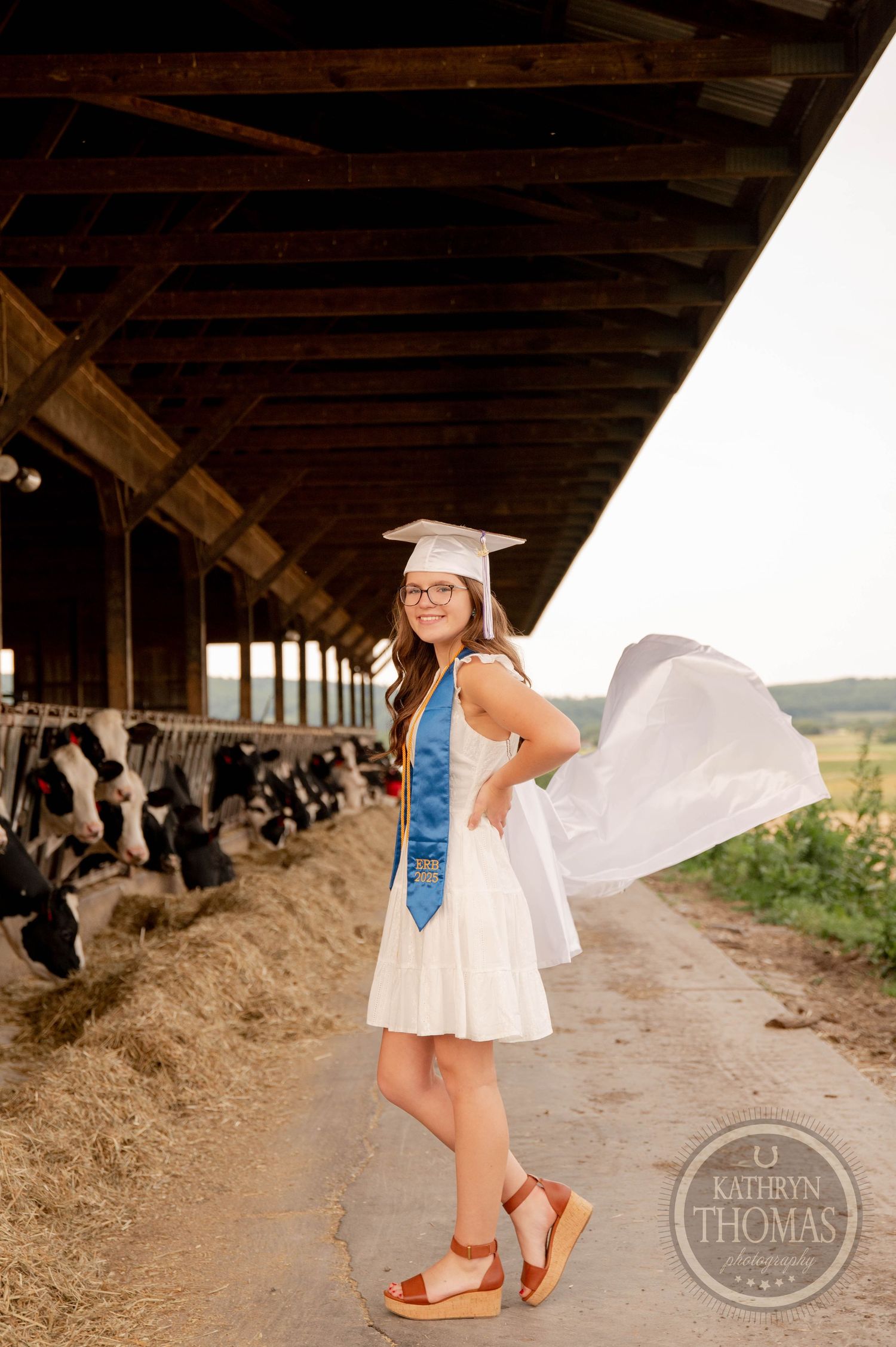 A graduate in a white dress and graduation cap poses in a barn aisle with dairy cows visible in the background.