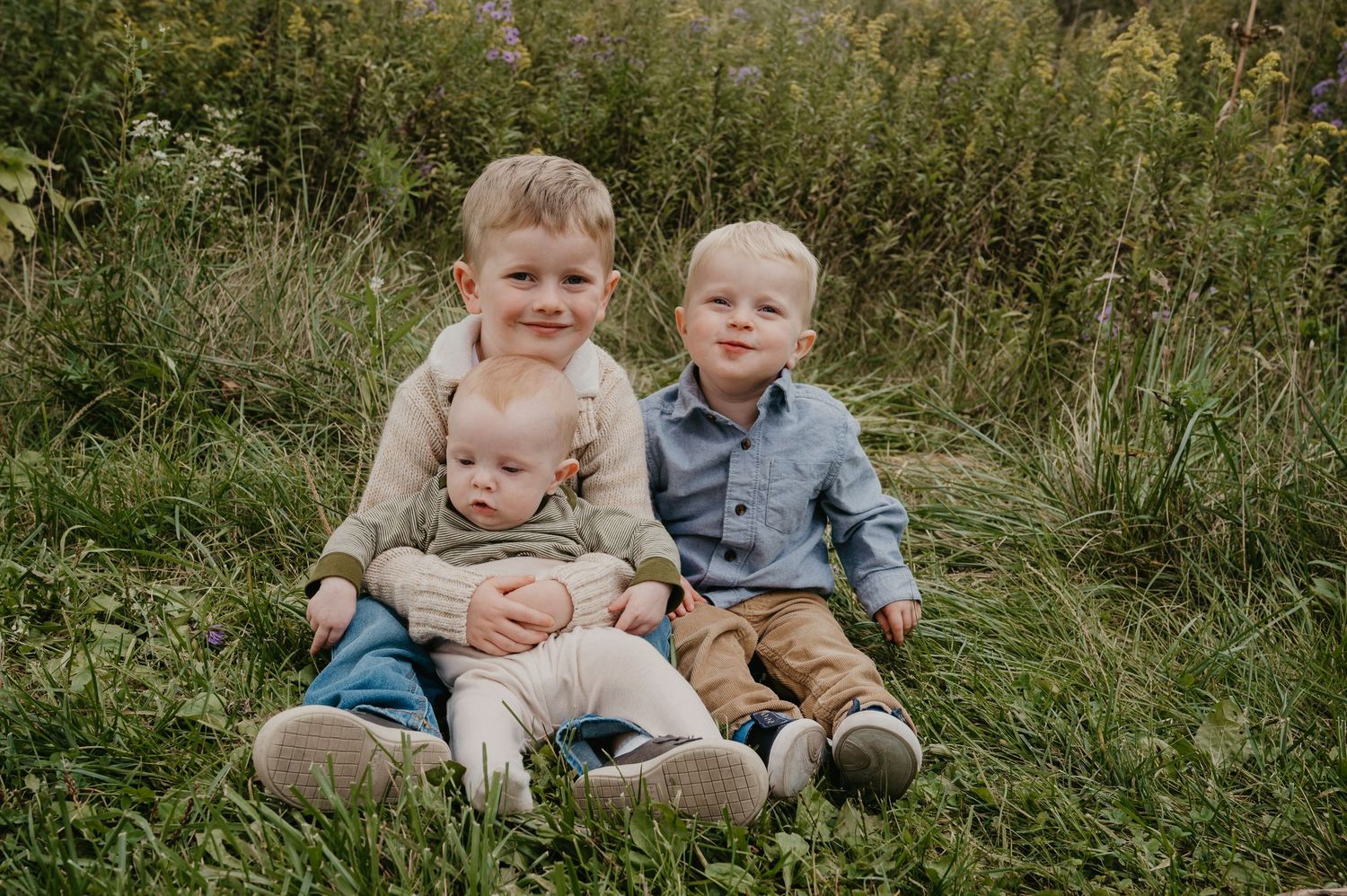 Three young brothers sitting together in a green meadow, oldest boy hugging younger siblings in a sweet outdoor portrait.