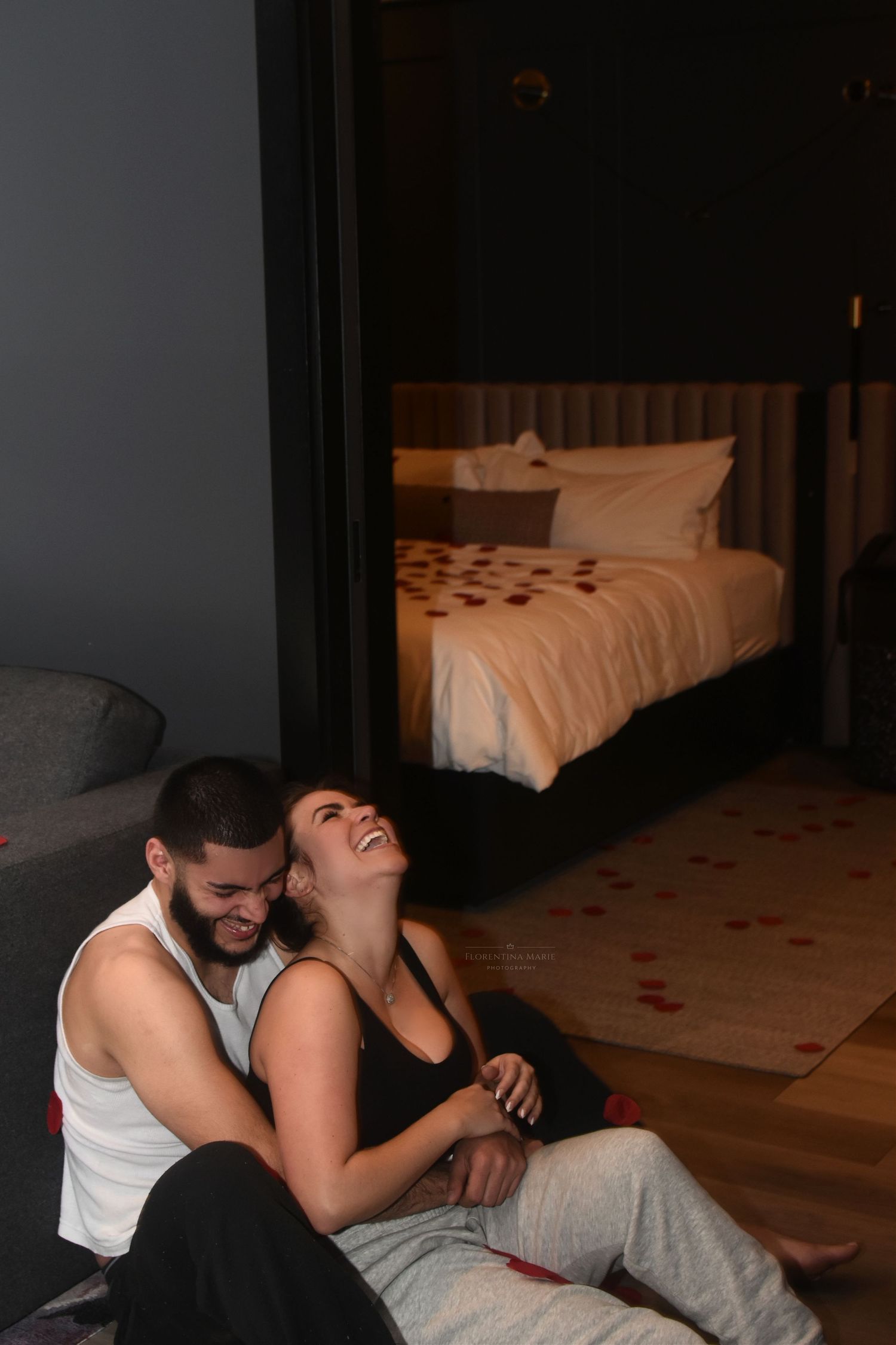 A couple shares an intimate moment of laughter while sitting on the floor of a dimly lit hotel room.