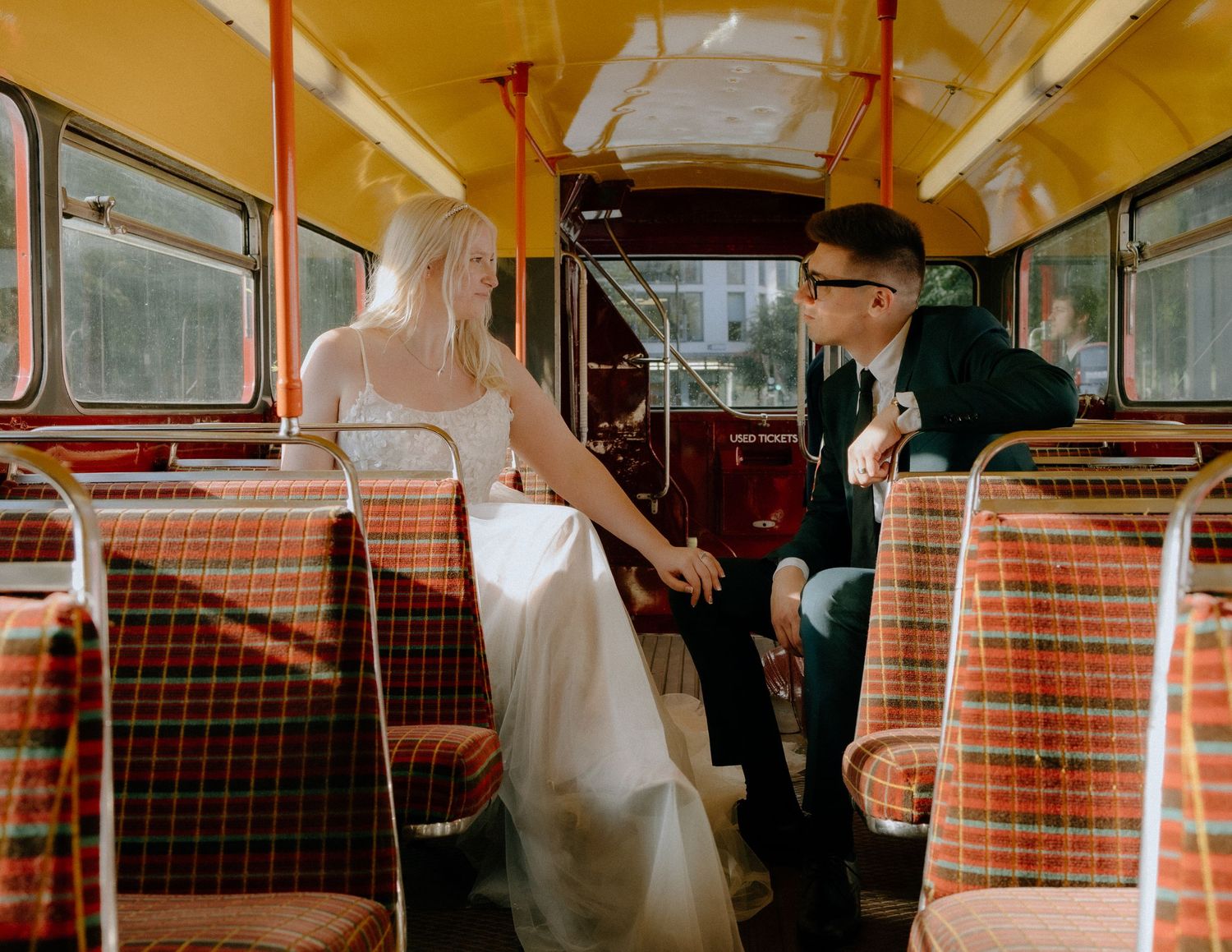 A couple in wedding attire sits inside a vintage bus with retro plaid seats.