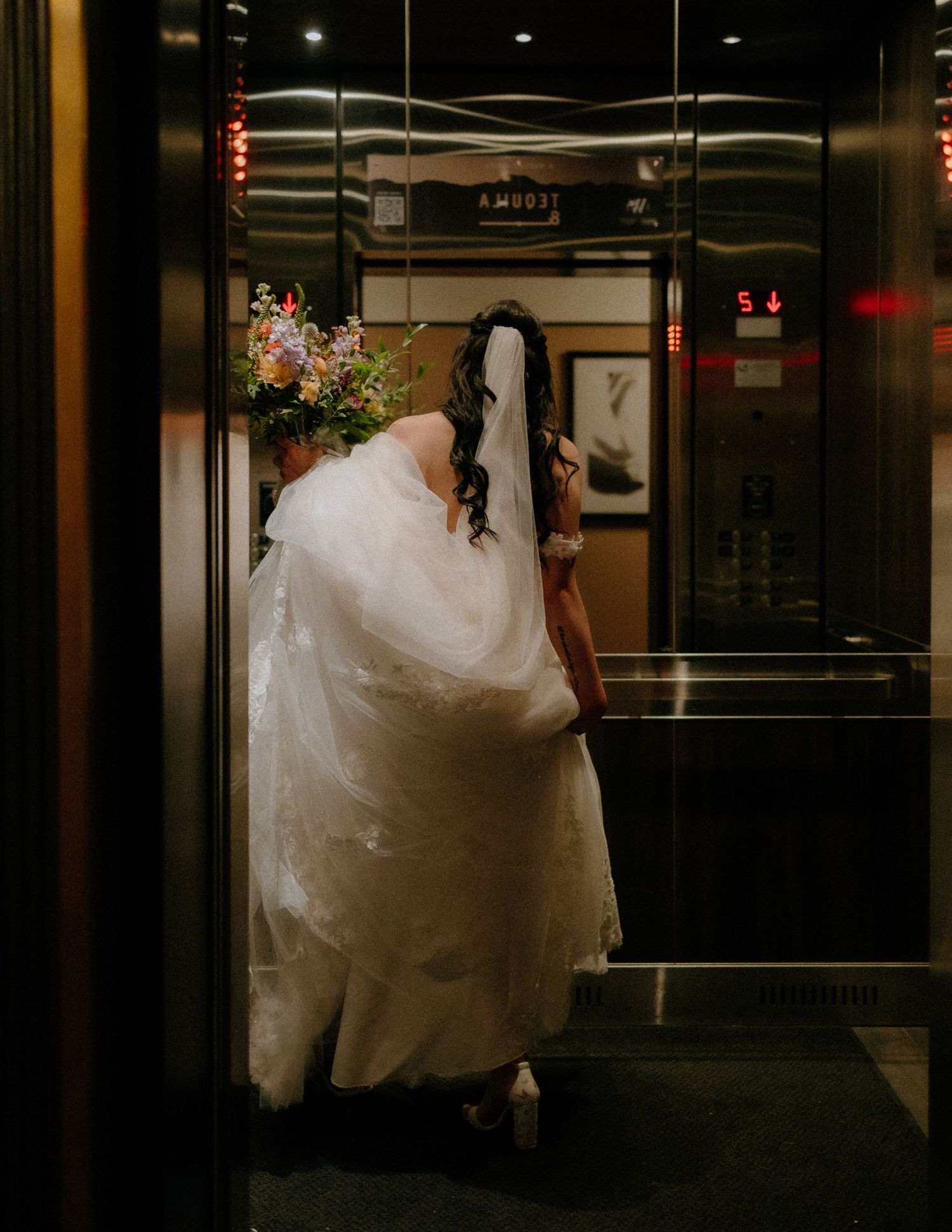 A bride in a flowing white dress steps into an elegant elevator holding a bouquet.