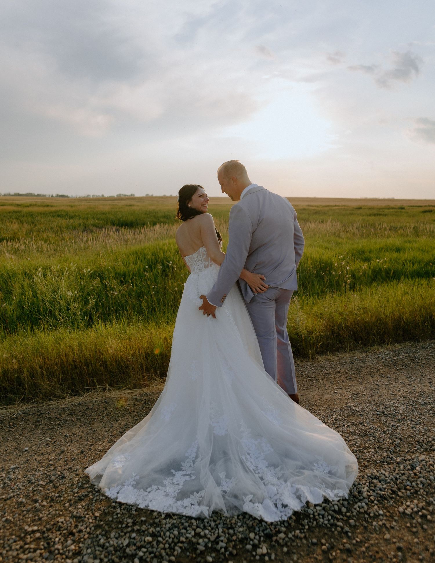 A romantic wedding couple embraces in a golden field at sunset with the bride wearing a flowing lace gown.