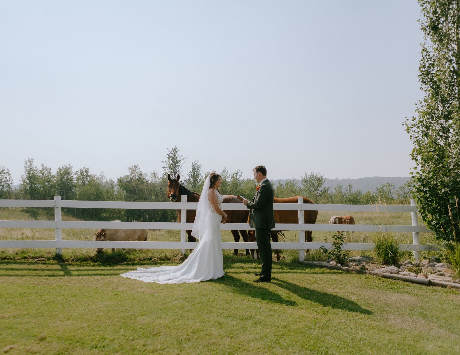 A wedding ceremony takes place by a white fence in a serene countryside setting.