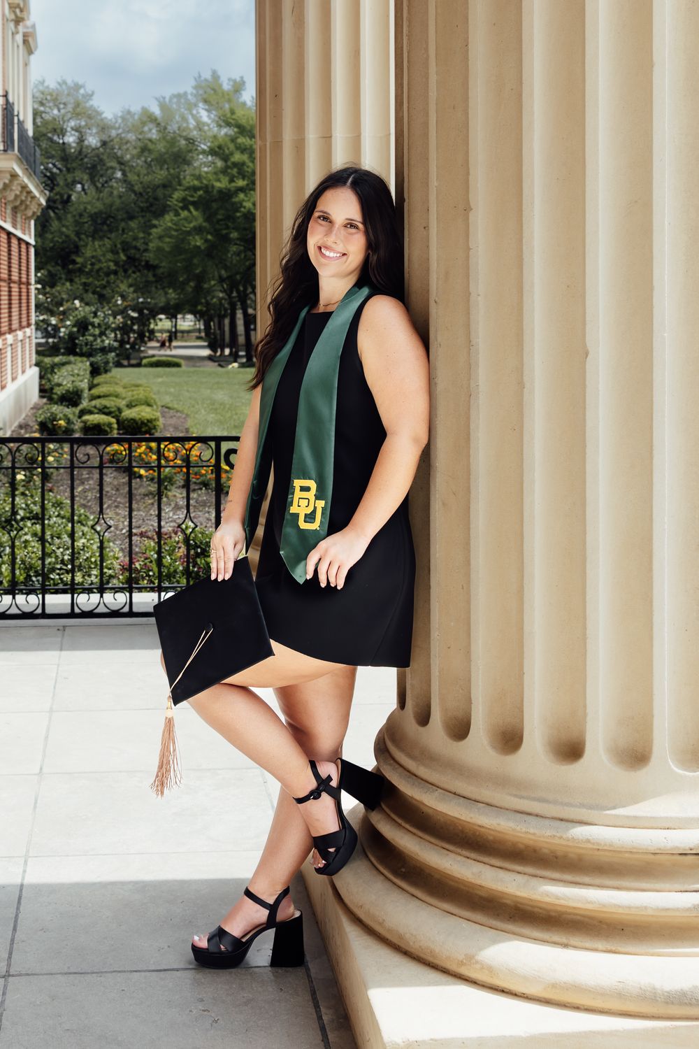 Baylor University graduate in black dress and green BU stole holds cap beside campus column.