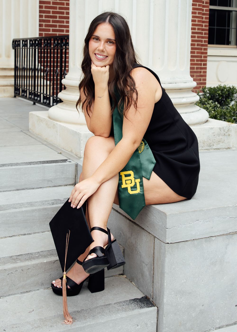 Young woman in black dress with Baylor University graduation sash and cap poses on brick steps outdoors.