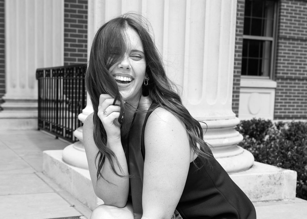 Black and white photo of a laughing young woman with long hair in a black dress posing by white columns.