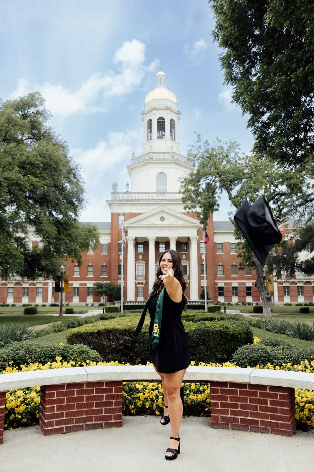 A graduate in a black dress tosses her cap in front of a brick university building with a white cupola.