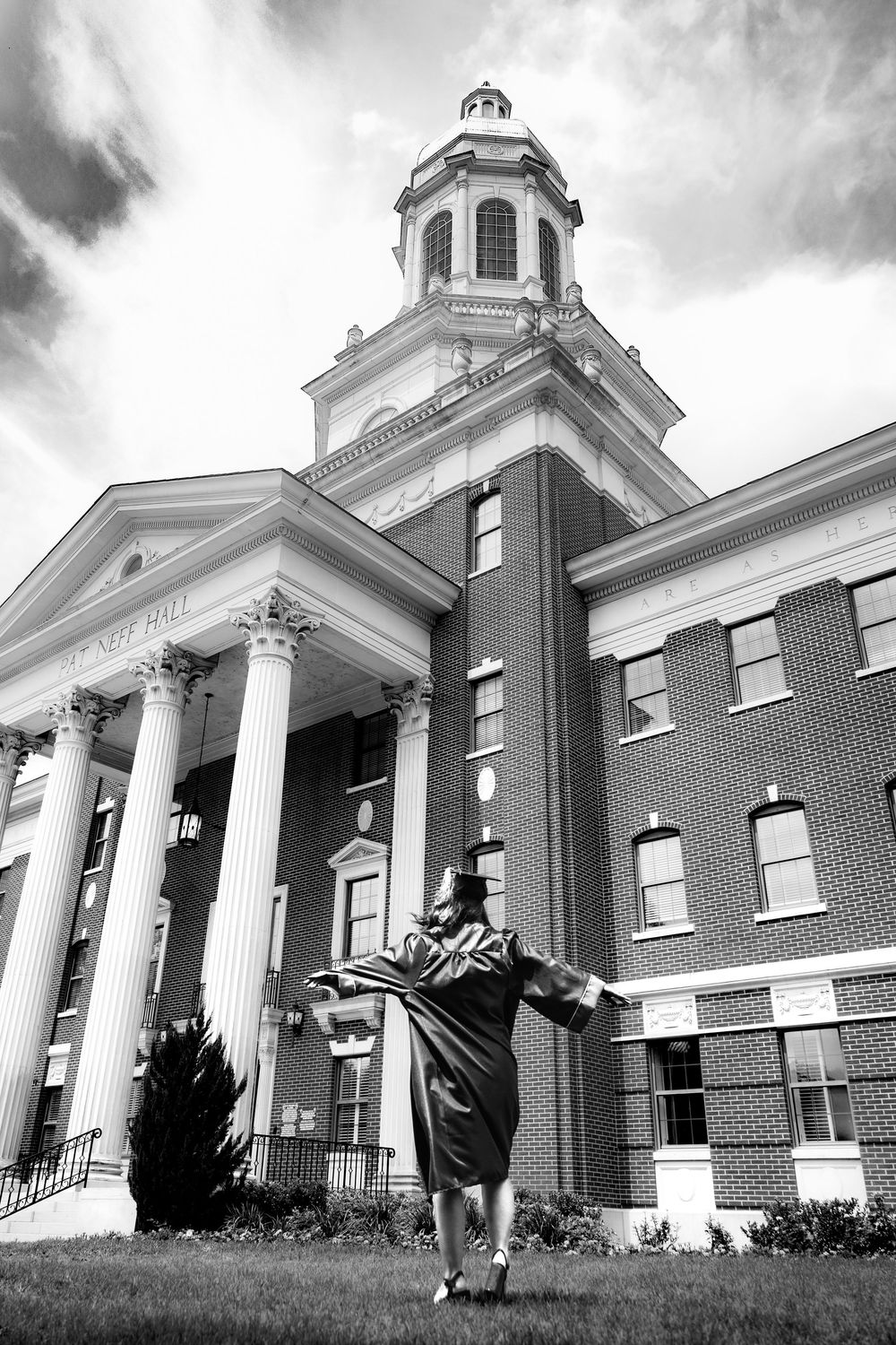 Graduate in cap and gown celebrates outdoors in front of a classic brick university building with white columns.