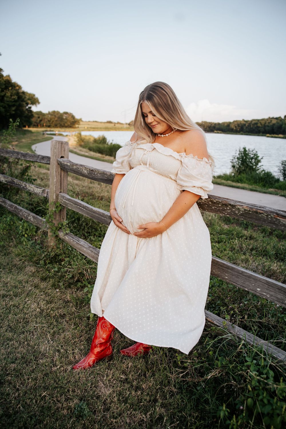 Pregnant woman in white off-shoulder dress and red cowboy boots holding belly near wooden fence by lake.