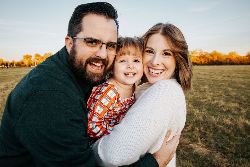 Happy family of three smiling outdoors in a sunny field during autumn, parents hugging young child in plaid shirt.