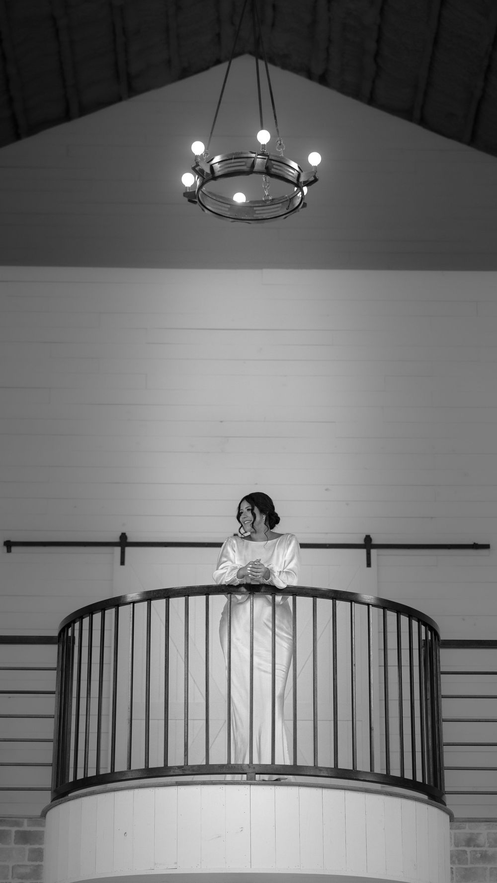Black and white photo of bride in white gown standing on balcony of rustic wedding venue chapel.