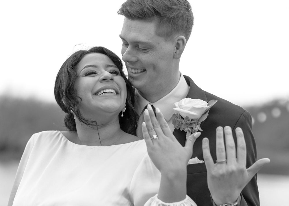 Black and white photo of smiling newlyweds showing off wedding rings near a lake.