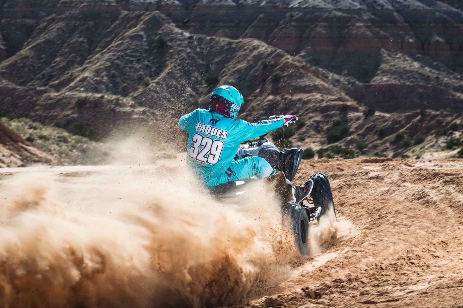 A dirt bike rider in turquoise gear kicks up dust while racing through rugged mountain terrain.