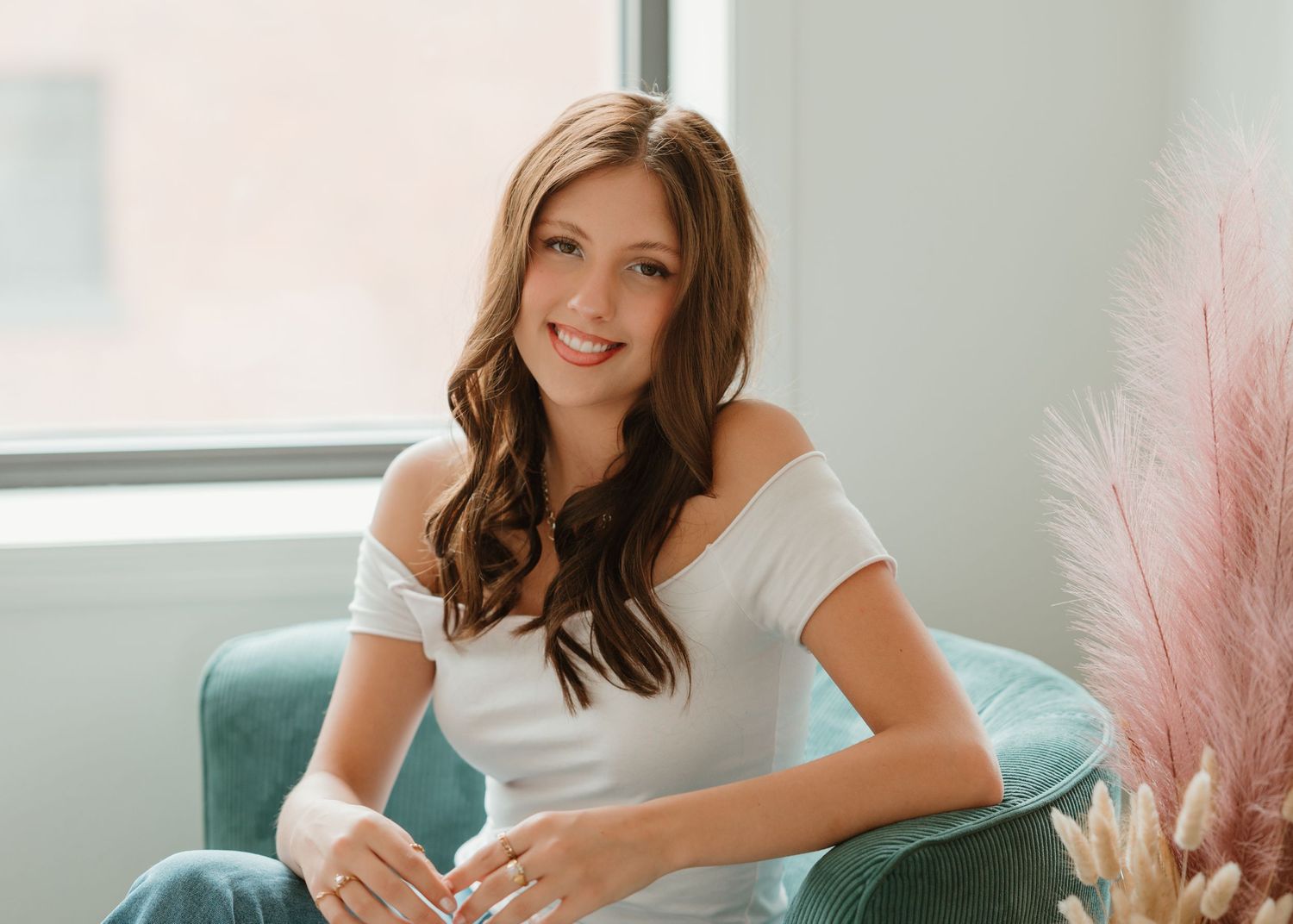Model in white off-shoulder top poses against teal chair and pink decor in natural light setting.