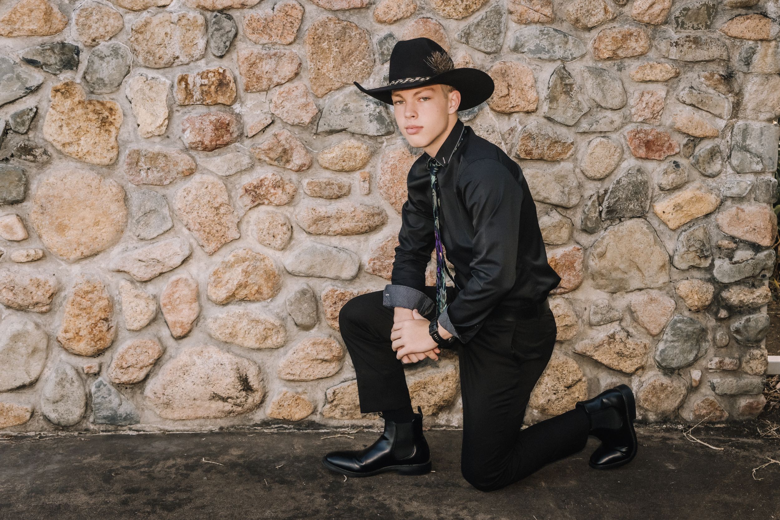 Western styled portrait of person in black attire kneeling against stone wall.