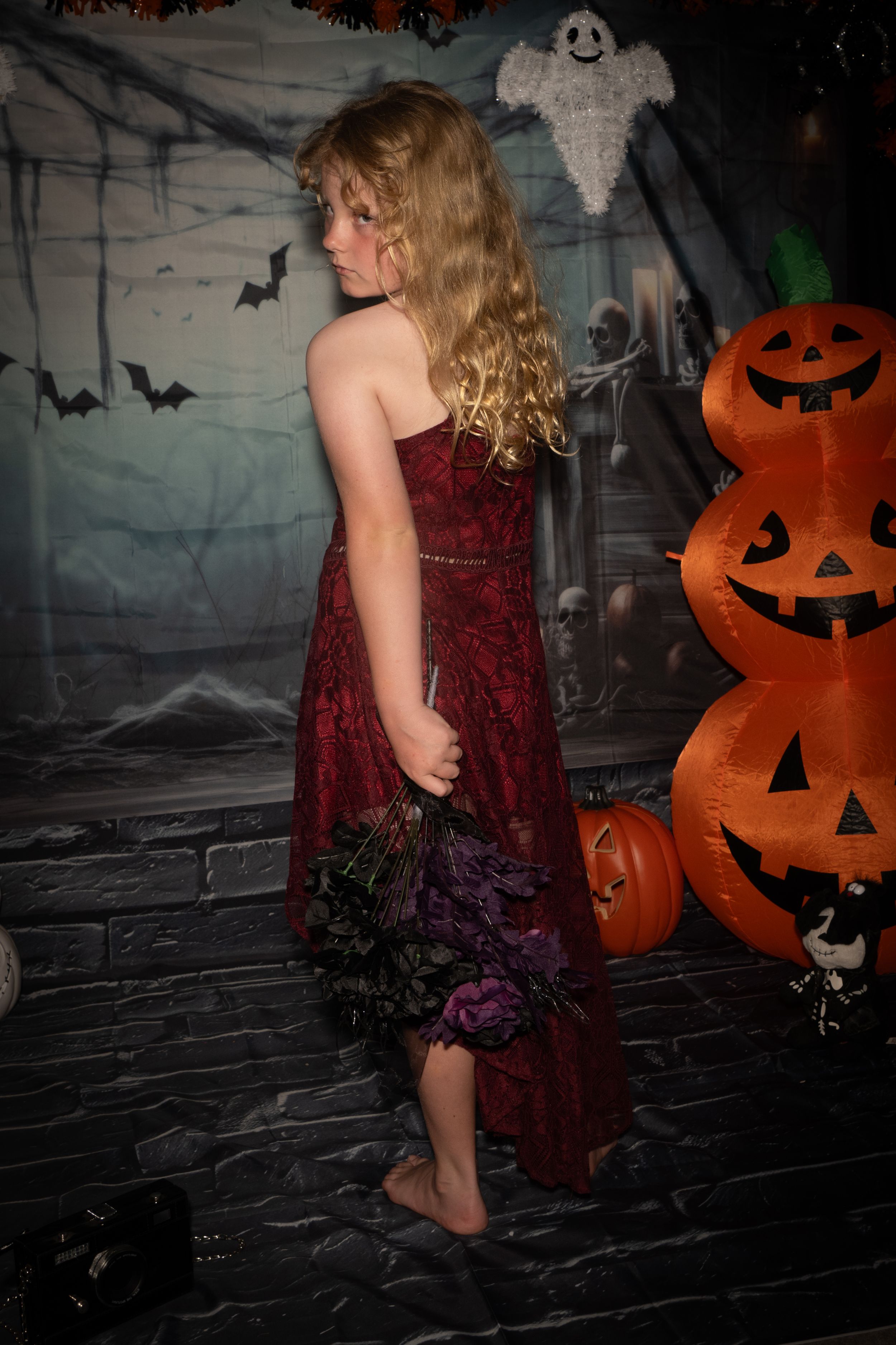 A red velvet dress stands out against a Halloween backdrop with stacked jack o'lanterns and spooky decorations.