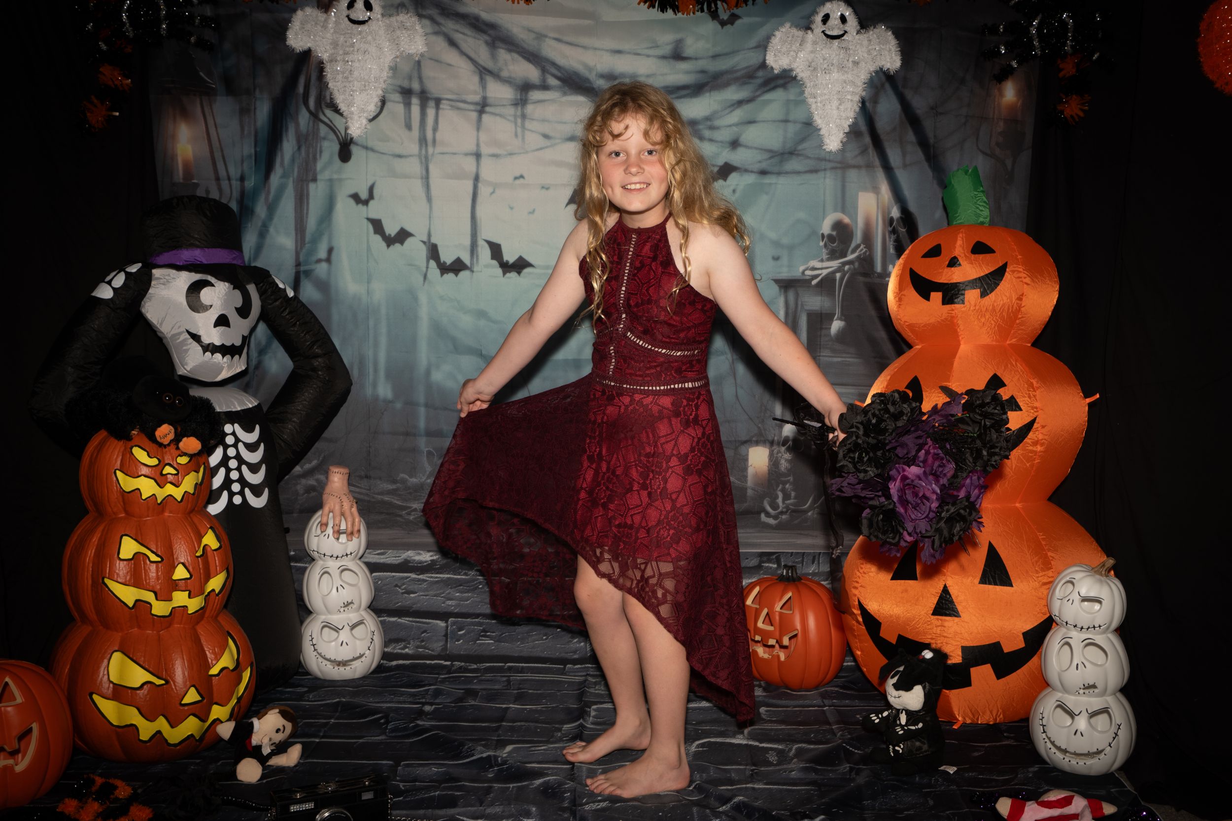 A person in a burgundy dress poses among Halloween decorations including jack-o-lanterns, ghosts and bats at a themed photoshoot.