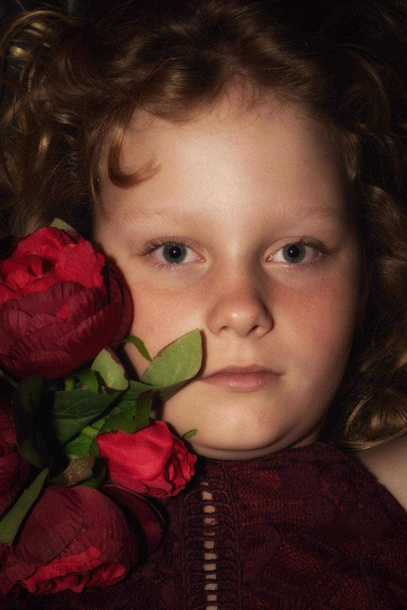 A close-up portrait with vibrant red roses against a dark background, wearing a burgundy top with soft lighting.