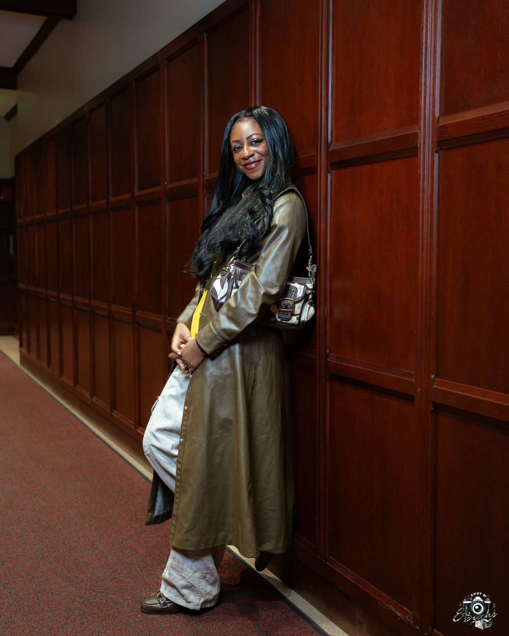 Someone wearing a long olive coat and baggy white pants poses casually against wooden paneled walls in a corridor.