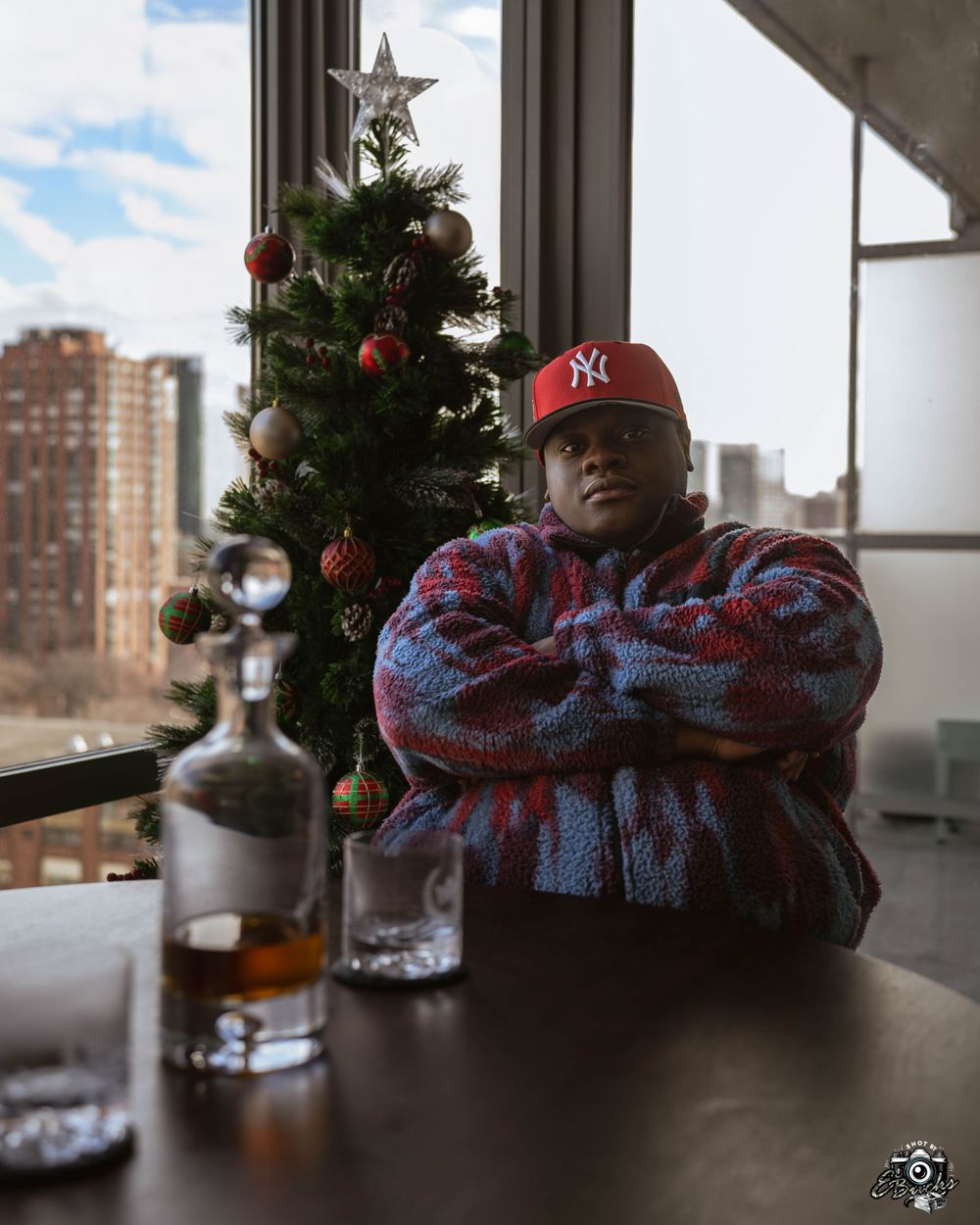 Person in Yankees hat and patterned jacket poses with arms crossed next to holiday decorations.