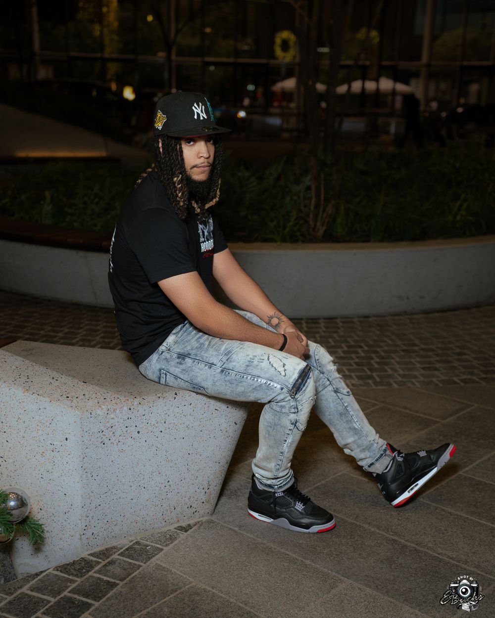 Person wearing a Yankees cap, black t-shirt and distressed jeans sits on a concrete ledge at night.