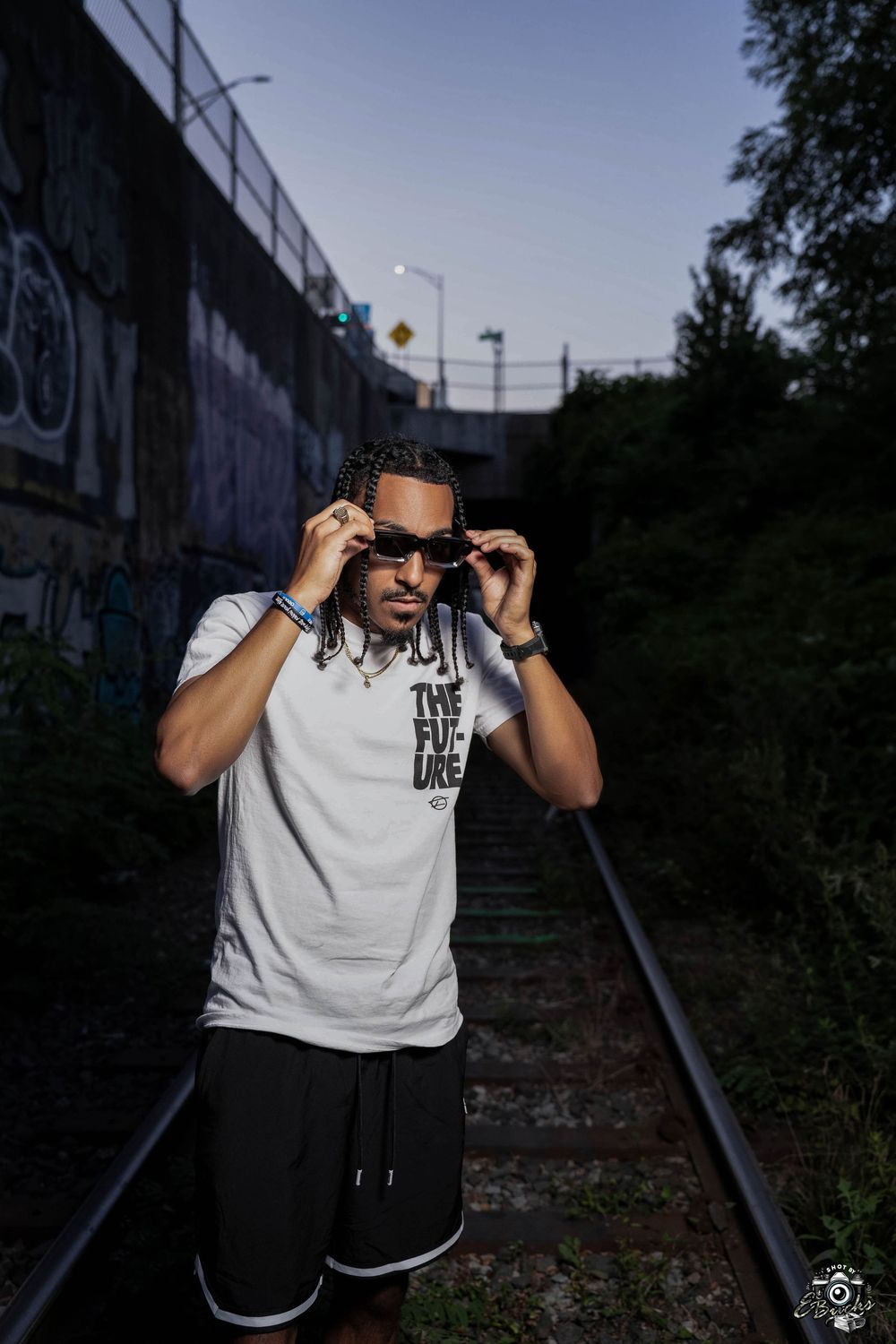 Athletic person in white t-shirt and shorts adjusting sunglasses while standing on train tracks at dusk.