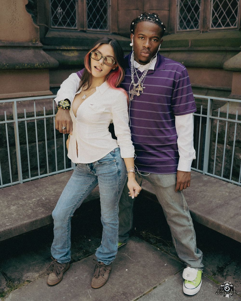 Two casual students pose against a stone building exterior with gothic windows at their university.