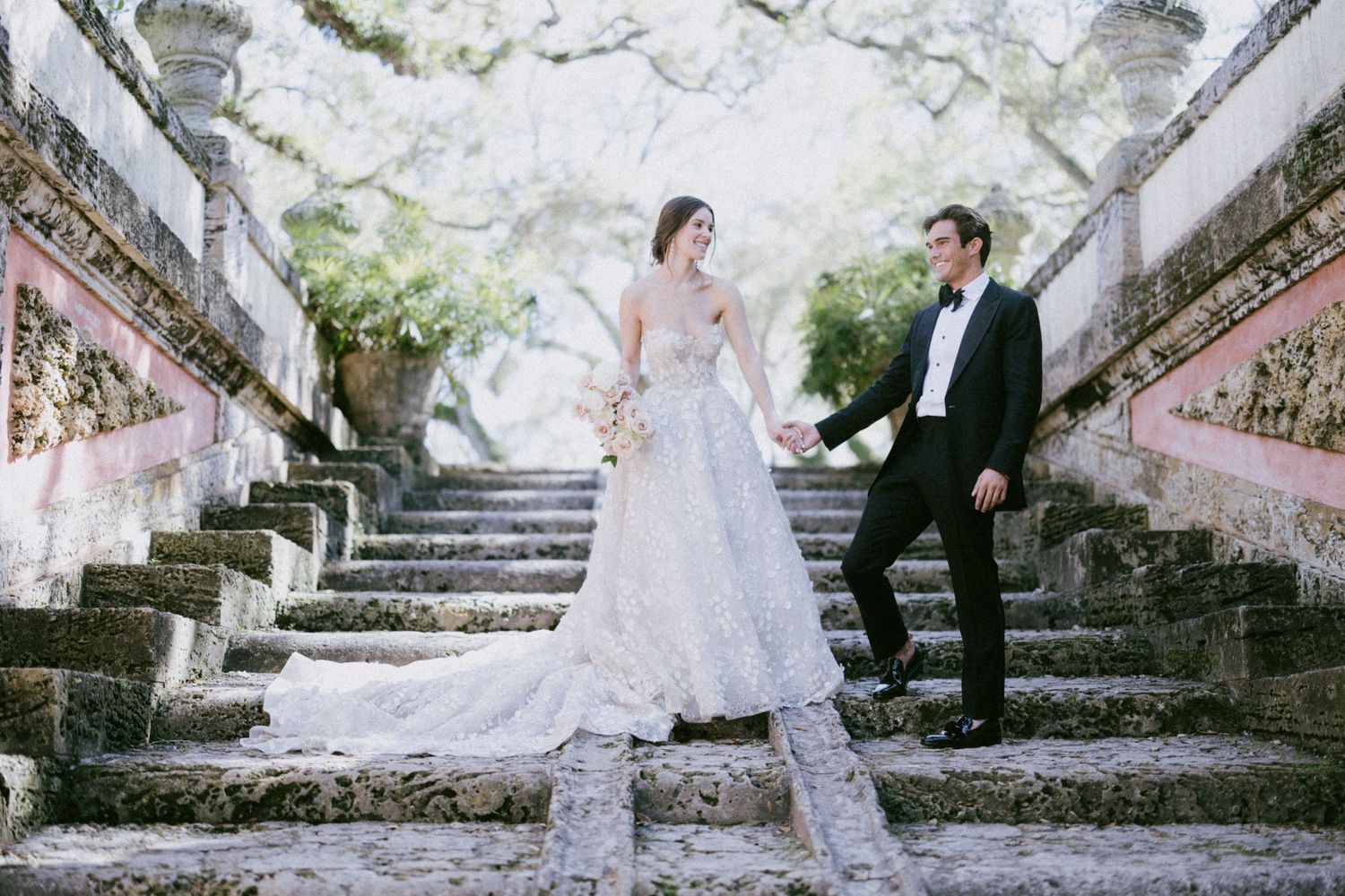 A couple in wedding attire poses on an ornate stone staircase surrounded by blossoming white trees.
