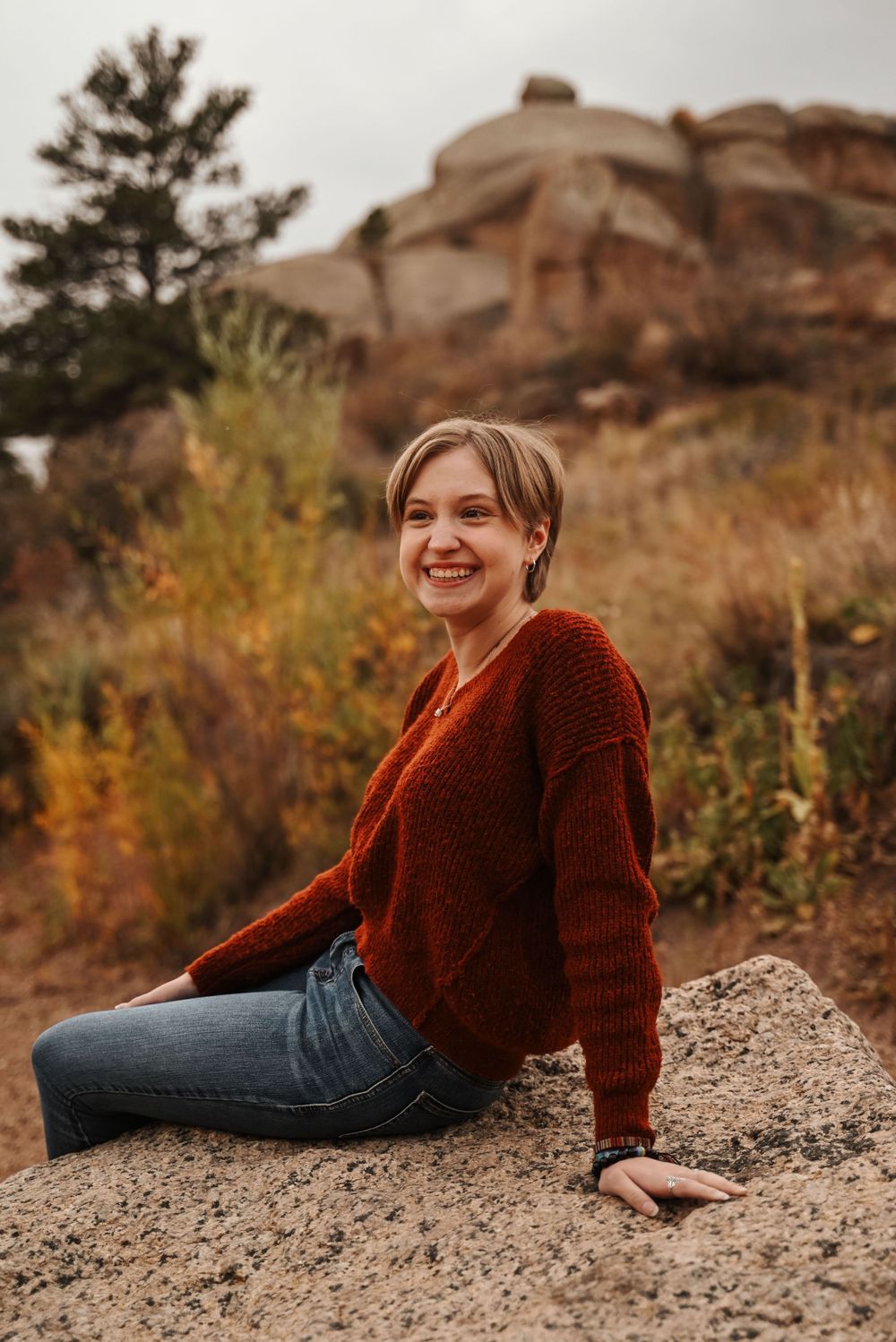 Person in red sweater sits on rocky outcrop against autumn foliage and desert rock formations.