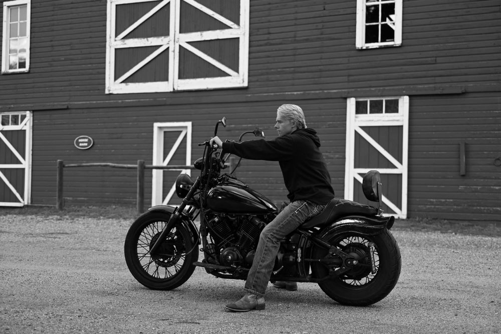 Black and white photo of person riding motorcycle in front of rustic barn building.