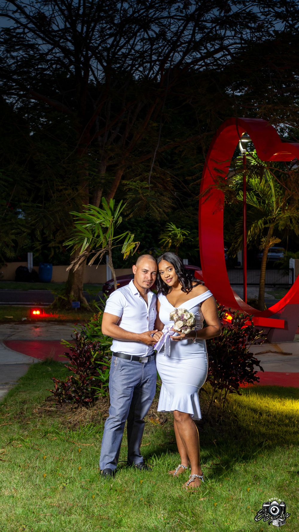 Couple in coordinated white outfits posing by red sculpture in garden at dusk.